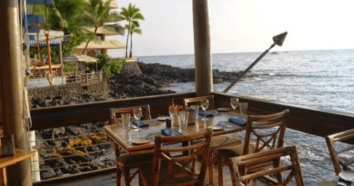 A seaside restaurant with wooden tables set for dining, overlooking the ocean and palm trees in the background.