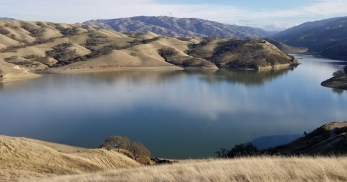 A serene landscape featuring a calm lake surrounded by rolling hills and distant mountains under a clear blue sky.
