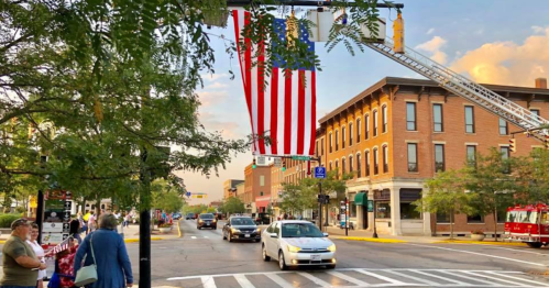 A street scene with a large American flag hanging above, surrounded by trees, buildings, and cars in a small town.