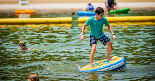 A child balances on a paddleboard in a calm lake, while another child swims nearby. Bright summer day.
