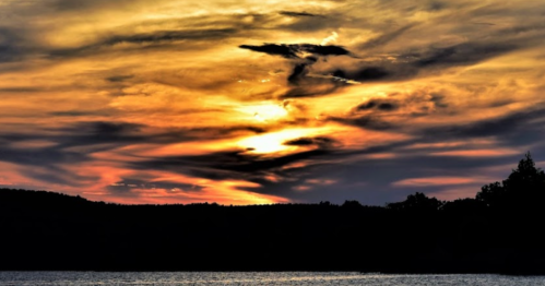 A vibrant sunset with swirling clouds in shades of orange, yellow, and purple over a calm lake and silhouette of trees.