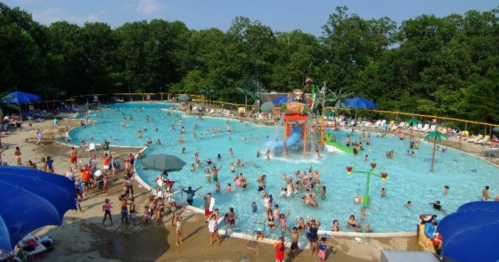 A busy outdoor pool with a water slide, surrounded by trees and umbrellas, filled with people enjoying the summer.