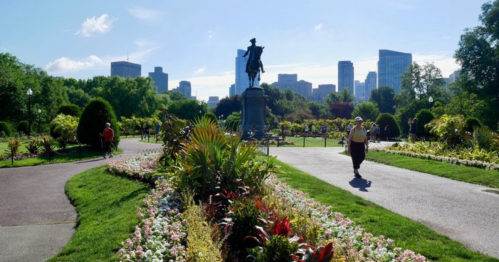 A park scene with a statue, colorful flower beds, and city skyscrapers in the background under a clear blue sky.