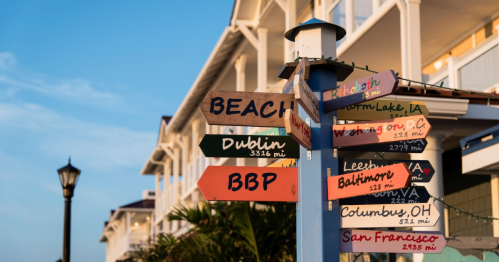 Colorful directional signs pointing to various cities and distances, with a beachside building in the background.