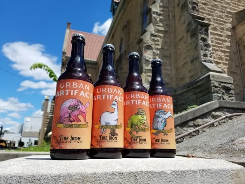 Four bottles of Urban Artifact beer with colorful labels featuring cartoon animals, set against a sunny outdoor backdrop.