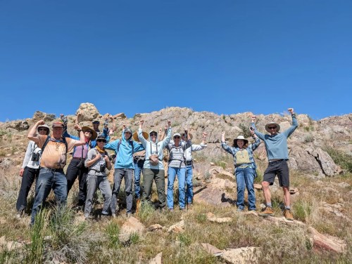 A group of hikers stands on a rocky hillside, cheering and raising their arms in celebration under a clear blue sky.