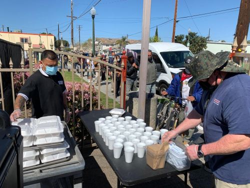 Two people serve food from a table with takeout containers and cups, while a line of people waits nearby.