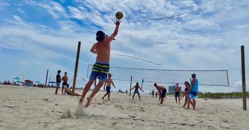 A group of people playing beach volleyball on a sunny day, with one player jumping to hit the ball.