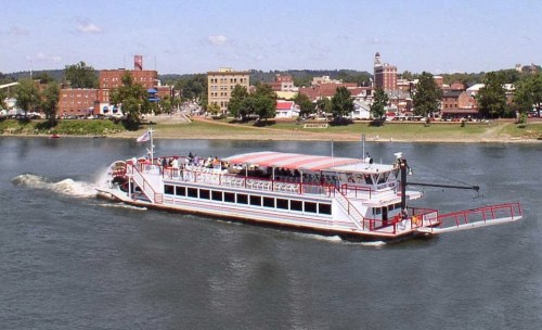 A riverboat with passengers cruises along a calm river, with a town and buildings visible in the background.