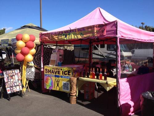 A colorful tent for Tutu's Shave Ice, featuring a menu sign, drinks, and balloons at a festive outdoor event.