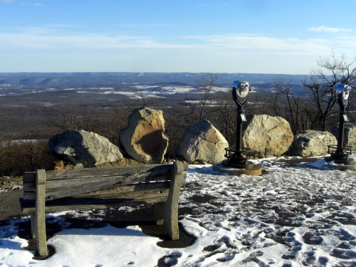 A scenic overlook with a bench, large rocks, and coin-operated binoculars, surrounded by snow-covered hills.