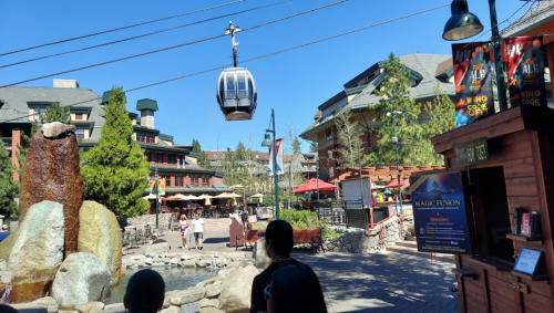 A scenic view of a resort area with a gondola, trees, and outdoor seating under a clear blue sky.