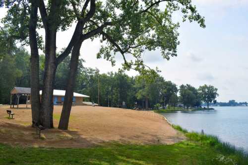 A sandy beach by a calm lake, with trees and a small building in the background under a cloudy sky.