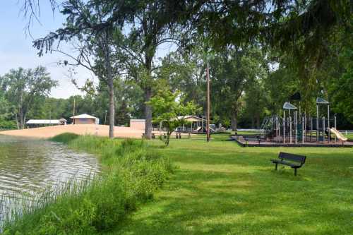 A serene park scene featuring a lake, grassy area, playground, and sandy beach under a clear blue sky.