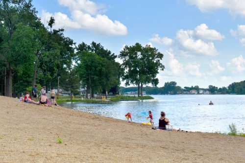 A sandy beach by a lake with families enjoying the water and trees in the background under a blue sky.