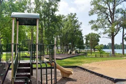 A playground with a slide and climbing structure, surrounded by trees and a lake in the background on a sunny day.