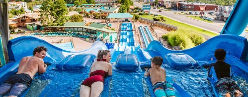 Aerial view of four people preparing to slide down a water slide at a water park, with colorful slides and greenery in the background.