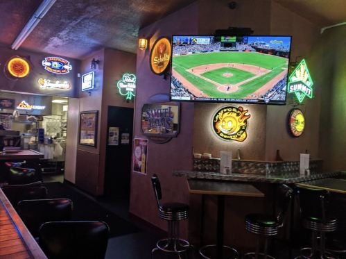 A cozy bar interior with neon signs, a large TV showing a baseball game, and empty stools at the counter.