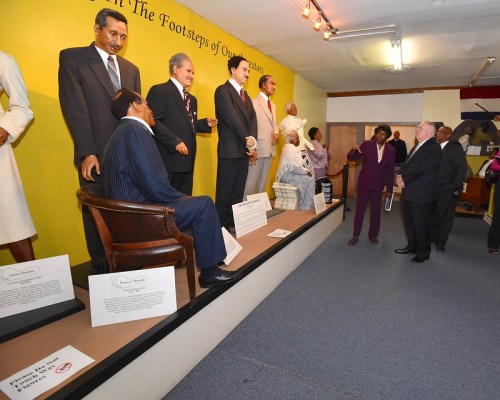 A group of life-sized figures in a museum exhibit, showcasing historical figures in formal attire. Visitors observe the display.