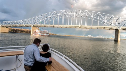 A couple sits on a boat, gazing at a large bridge and a riverboat under a cloudy sky.