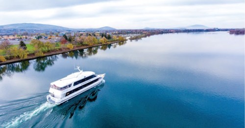 A white boat glides through calm waters, surrounded by trees and distant hills under a cloudy sky.
