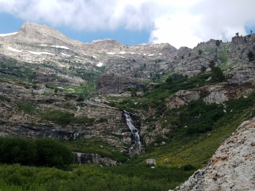 A scenic mountain landscape featuring a waterfall cascading down rocky terrain, surrounded by lush greenery and clouds.
