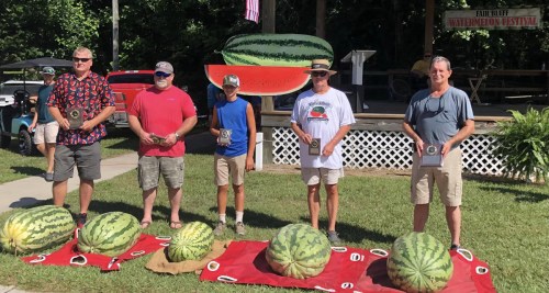 Five men stand with trophies in front of large watermelons at a watermelon festival, with a decorated stage in the background.