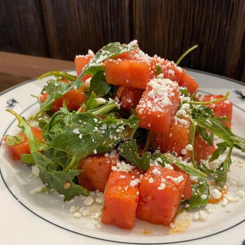 A vibrant salad of diced watermelon, arugula, and crumbled cheese, served on a white plate with a decorative rim.