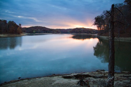 A serene lake at sunset, surrounded by trees and hills, reflecting colorful clouds in the calm water.