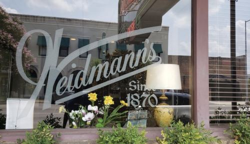 Window display featuring "Heidmann's Since 1870" sign, with plants and a lamp visible inside.