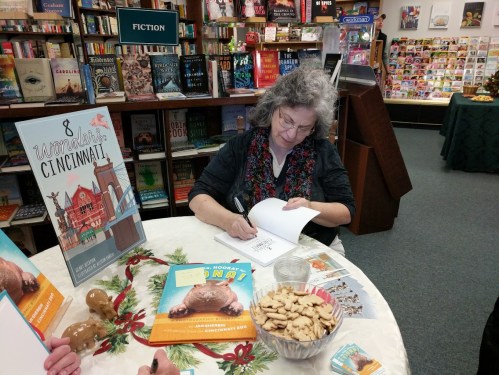 A woman signs a book at a table with cookies, surrounded by bookshelves in a cozy bookstore setting.