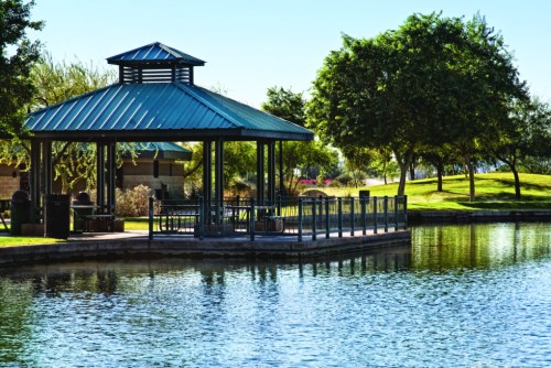 A serene park scene featuring a gazebo by a calm lake, surrounded by lush greenery and clear blue skies.