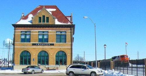 Historic train station in Creston, with snow on the roof and a train passing by in the background. Clear blue sky.