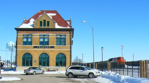 Historic Creston train station with snow-covered ground, parked cars, and a train in the background under a clear blue sky.