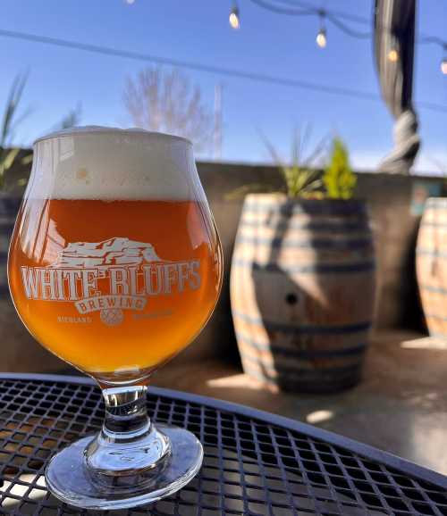 A glass of amber beer with foam sits on a table, with barrels and a clear sky in the background.