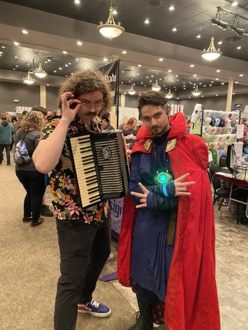 Two men pose in a convention setting; one plays an accordion, while the other wears a superhero costume with a cape.