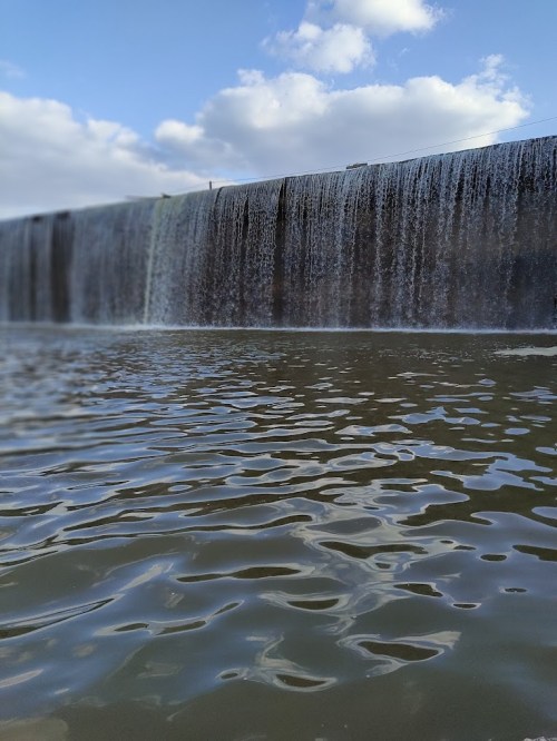 A large waterfall cascading over a stone wall into a calm body of water, under a blue sky with scattered clouds.