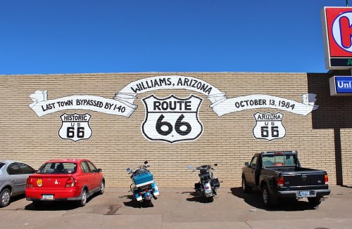 Mural in Williams, Arizona, celebrating Route 66 with text and parked cars below. Clear blue sky above.