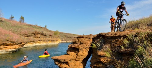 A cyclist navigates rocky terrain near a river, while two kayakers paddle in the water under a clear blue sky.