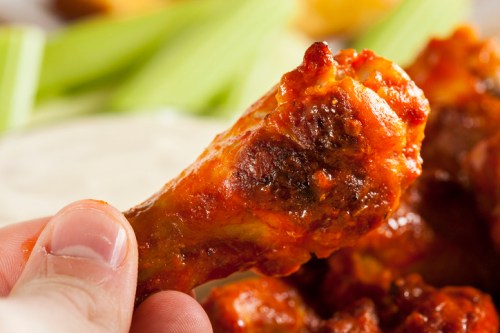 A close-up of a hand holding a spicy chicken wing, with celery sticks and dipping sauce in the background.