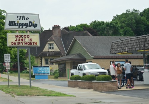 A sign for The Whippy Dip announces Dairy Month, with people gathered outside and a white truck parked nearby.