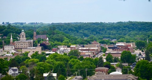 A panoramic view of a small town surrounded by lush greenery, featuring historic buildings and a tree-lined landscape.