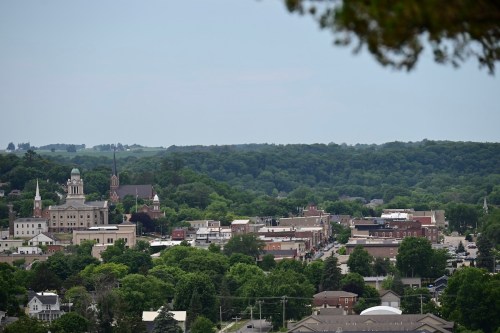 A panoramic view of a small town surrounded by green hills, featuring historic buildings and tree-lined streets.
