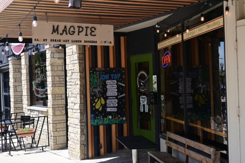 Exterior of a café named "Magpie," featuring a sign, outdoor seating, and a colorful menu board.