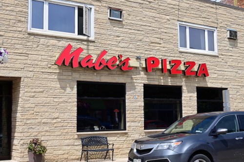 Exterior of Mabe's Pizza, featuring a stone facade and a bright red sign with the restaurant's name. A parked car is visible.