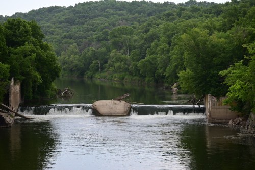 A serene river scene with a small dam, surrounded by lush green trees and hills in the background.