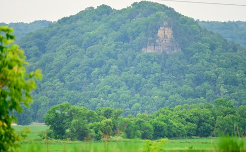 A lush green hillside with a rocky outcrop, surrounded by trees and open fields under a cloudy sky.