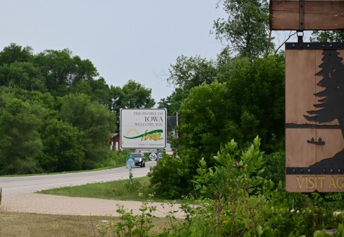 A welcome sign for Iowa along a road, surrounded by greenery and trees.