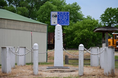 A historical marker with a stone monument surrounded by chains, located near a building and trees.