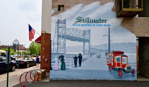 A mural depicting Stillwater, featuring a bridge, vintage car, and people in historical attire, with an American flag nearby.